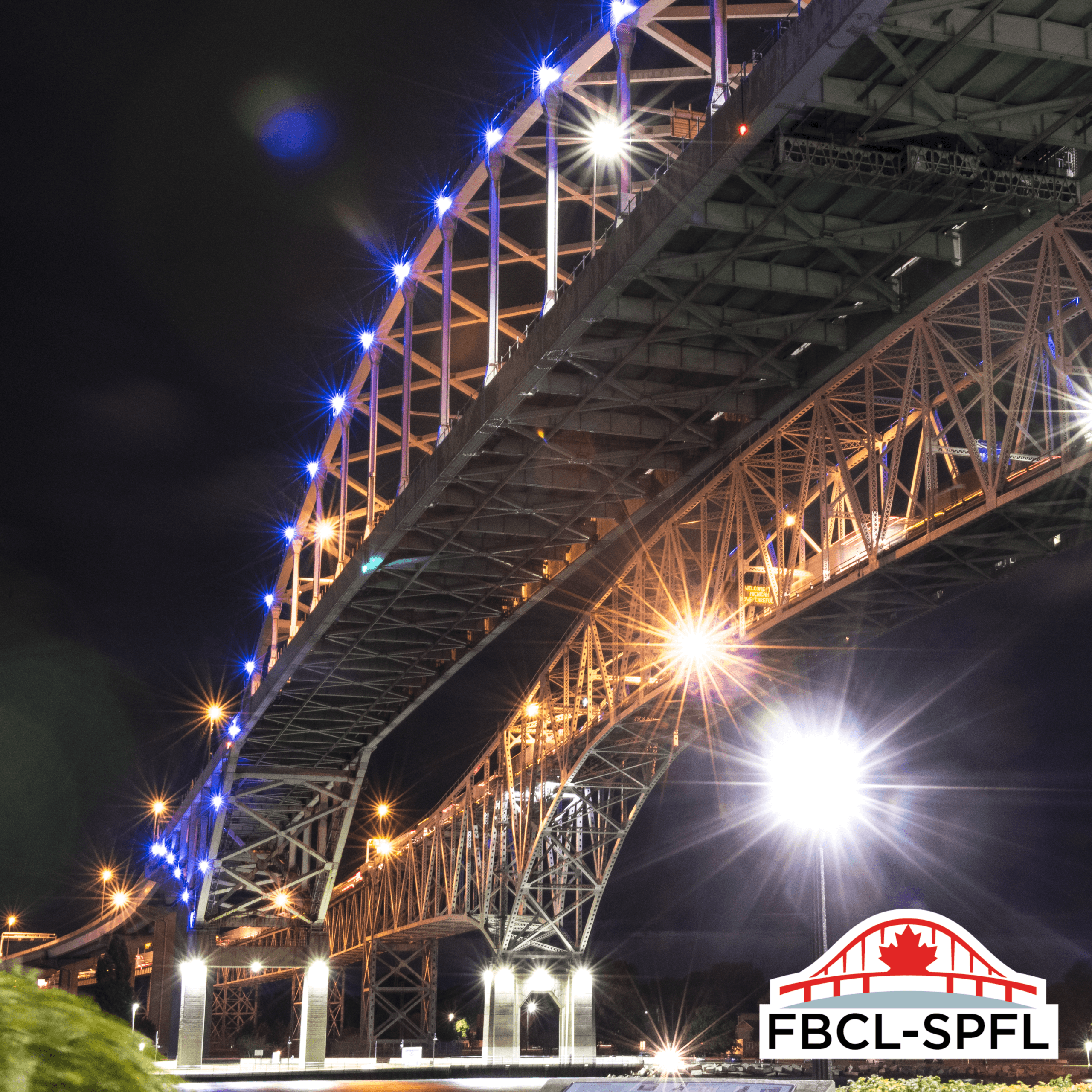 Low-angle nighttime view beneath both spans of the Blue Water Bridge, with blue LED lights illuminating the upper span and warm white lights illuminating the span below. The FBCL logo appears in the lower-right corner.